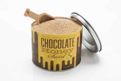 An open tin of Beautiful Briny Sea's Chocolate Honey Swirl Sugar, filled with chocolate honey-infused organic cane sugar and a small wooden scoop, with the lid resting nearby.