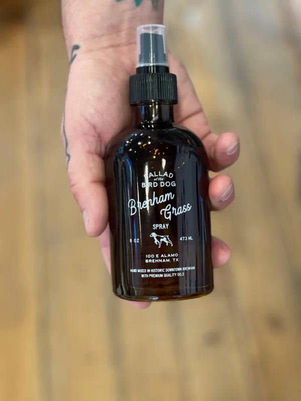 A hand holds a brown glass Dog Spray by Ballad of the Bird Dog, 16 oz, labeled "Ballad of the Bird Dog," set against wood flooring.