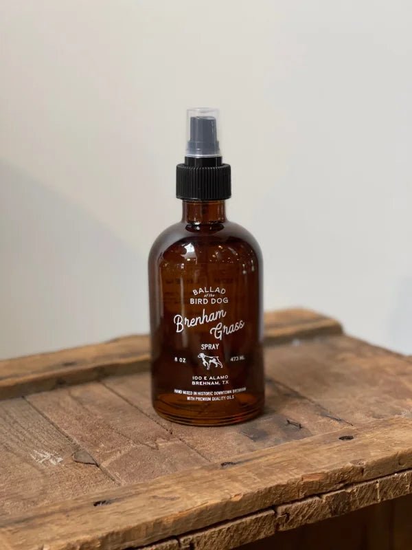A brown glass spray bottle, labeled "Dog Spray | Ballad of the Bird Dog" from Ballad of the Bird Dog, sits on a rustic wooden surface against a plain background.