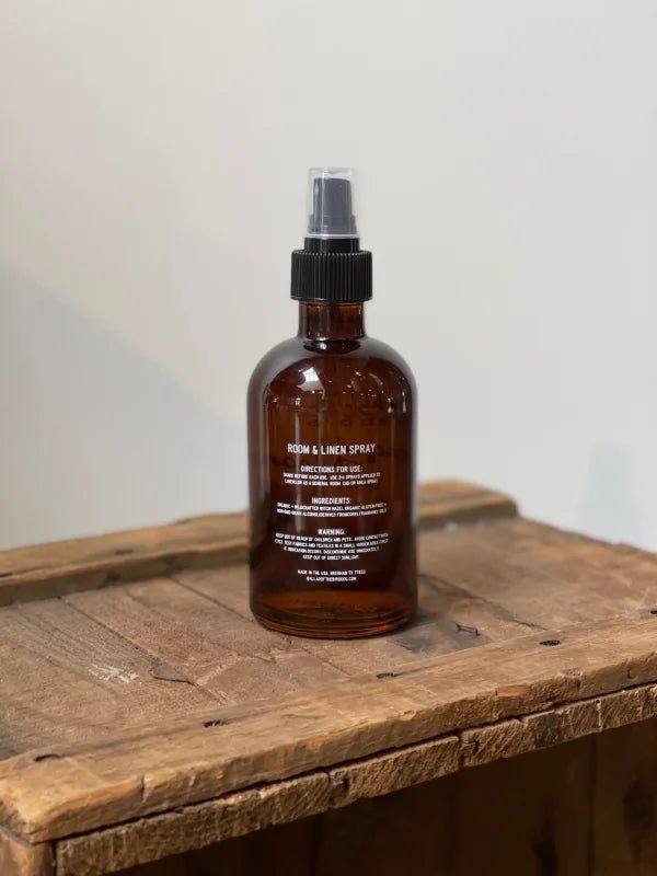 A brown glass bottle labeled "Dog Spray | Ballad of the Bird Dog" by Ballad of the Bird Dog, infused with witch hazel and essential oils, features a black spray nozzle and rests on a rustic wooden surface.