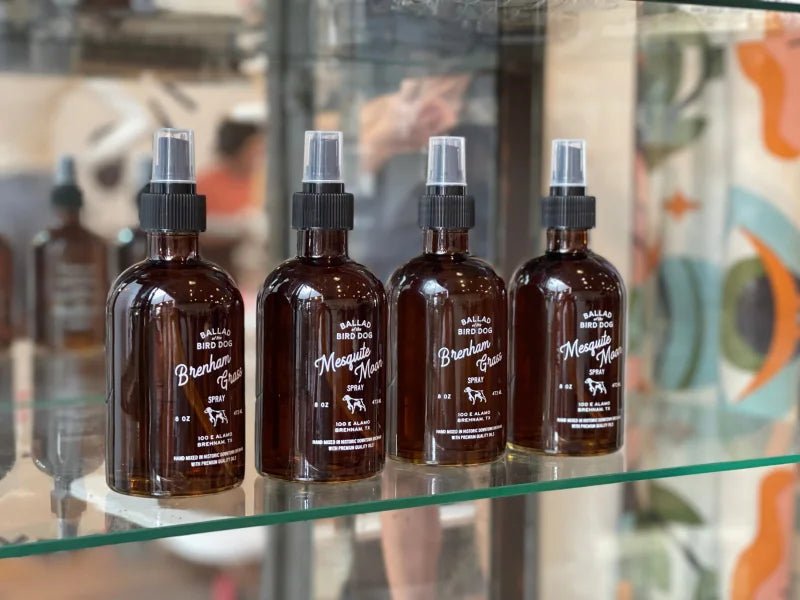 Four brown spray bottles of Ballad of the Bird Dog Dog Spray—formulated with witch hazel and essential oils—are lined up on a glass shelf against a colorful, blurred background.