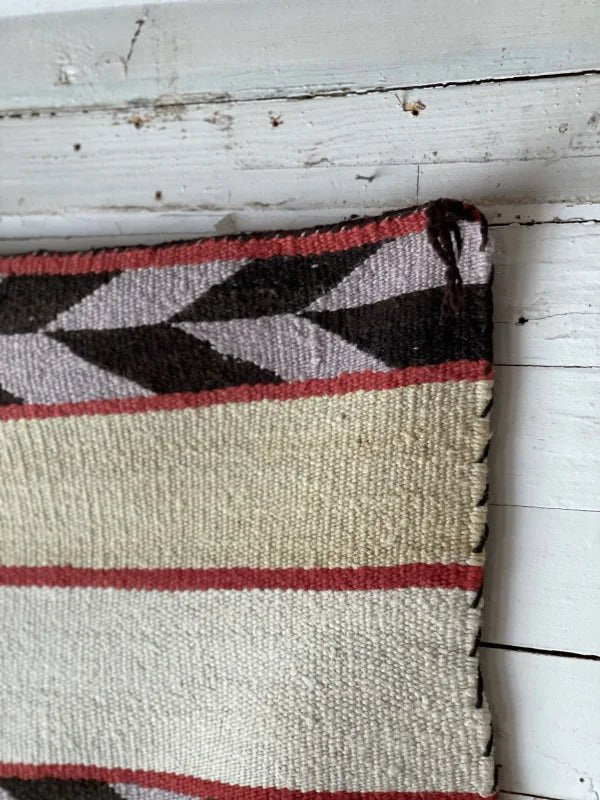 A close-up of the Vintage Double Saddle Blanket Circa 1940, featuring black, white, and red geometric patterns, is shown on a worn white wooden floor.