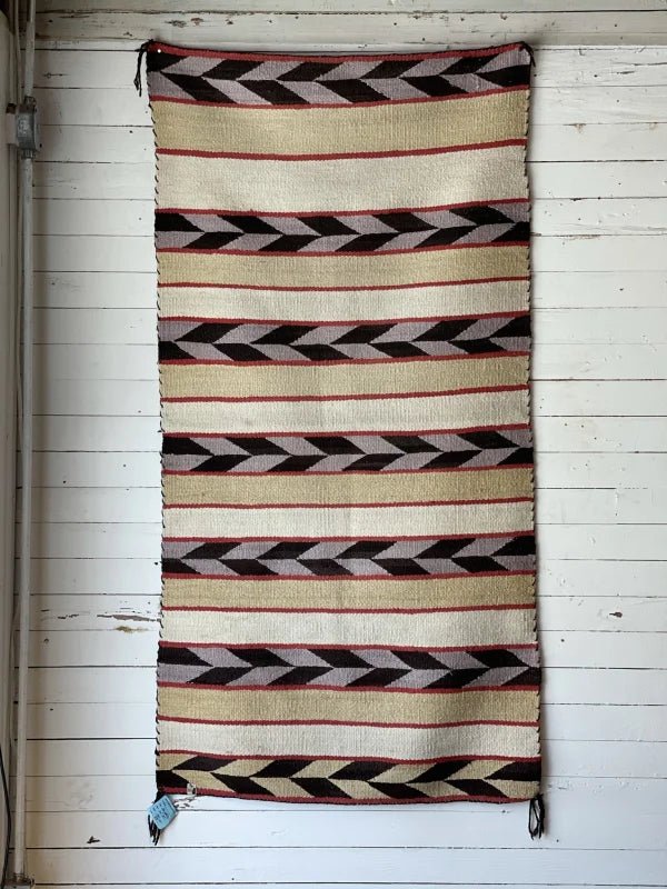 The Vintage Double Saddle Blanket Circa 1940, a rectangular woven rug with black, brown, red, and beige geometric patterns, hangs on a white wooden wall.
