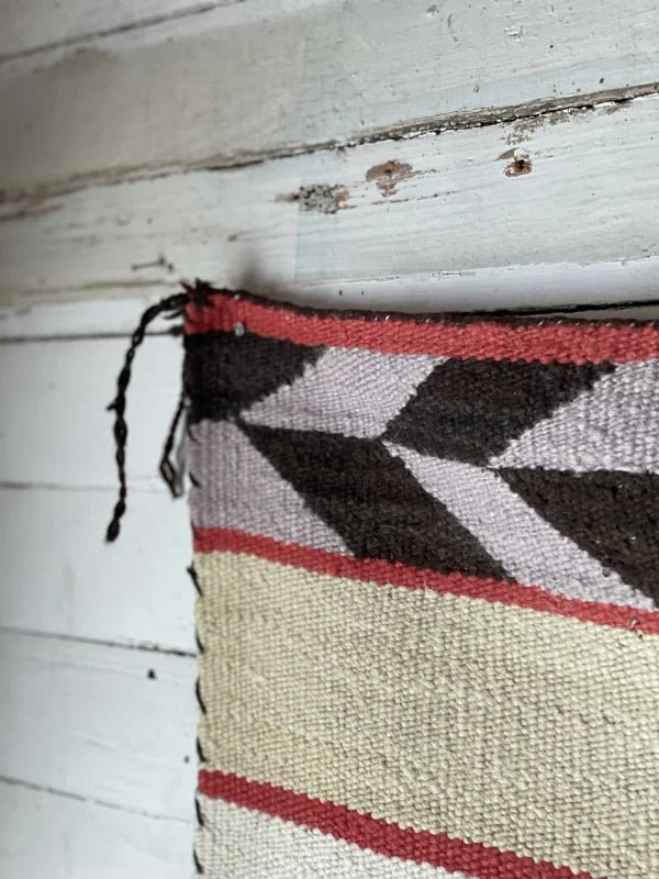 A close-up of the Double Saddle Blanket Circa 1940 by Vintage, featuring red, black, and cream geometric arrow feather patterns, displayed on a weathered white wooden wall.