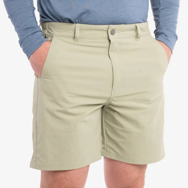 A person wears Duck Camp Drifter Shorts—quick-dry khaki fishing shorts—with hands in pockets and a blue long-sleeve shirt, shown waist to mid-thigh against a plain background.