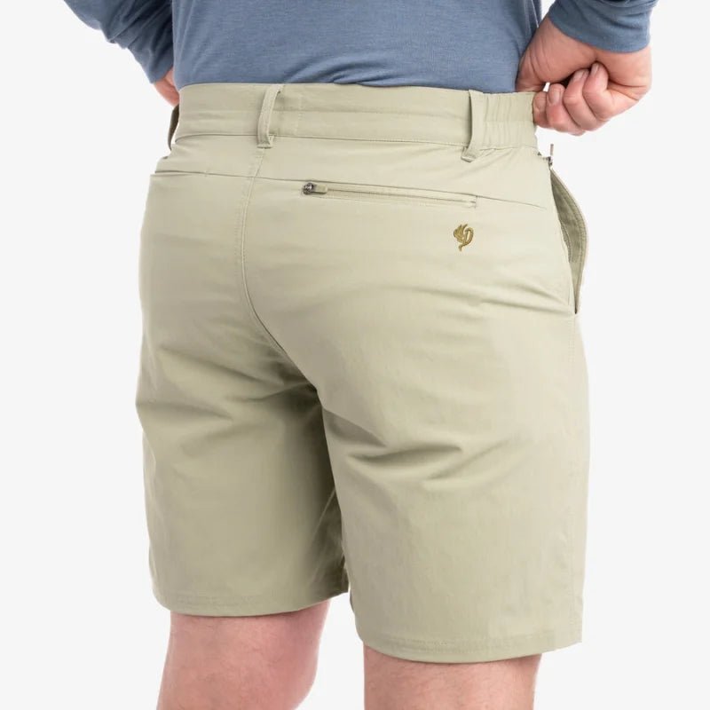 A person shown from the waist down, viewed from behind, wears Duck Camp Drifter Shorts in light khaki with UPF40+ protection, a back zipper pocket, and a small embroidered logo.