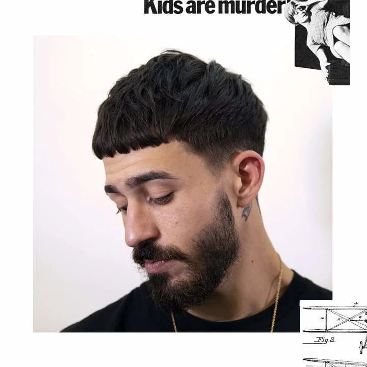 A man with a dark beard and short, textured hair looks down, showcasing effortless styling and a natural matte finish achieved using Uppercut Deluxe Easy Hold; background features partial text and graphics.