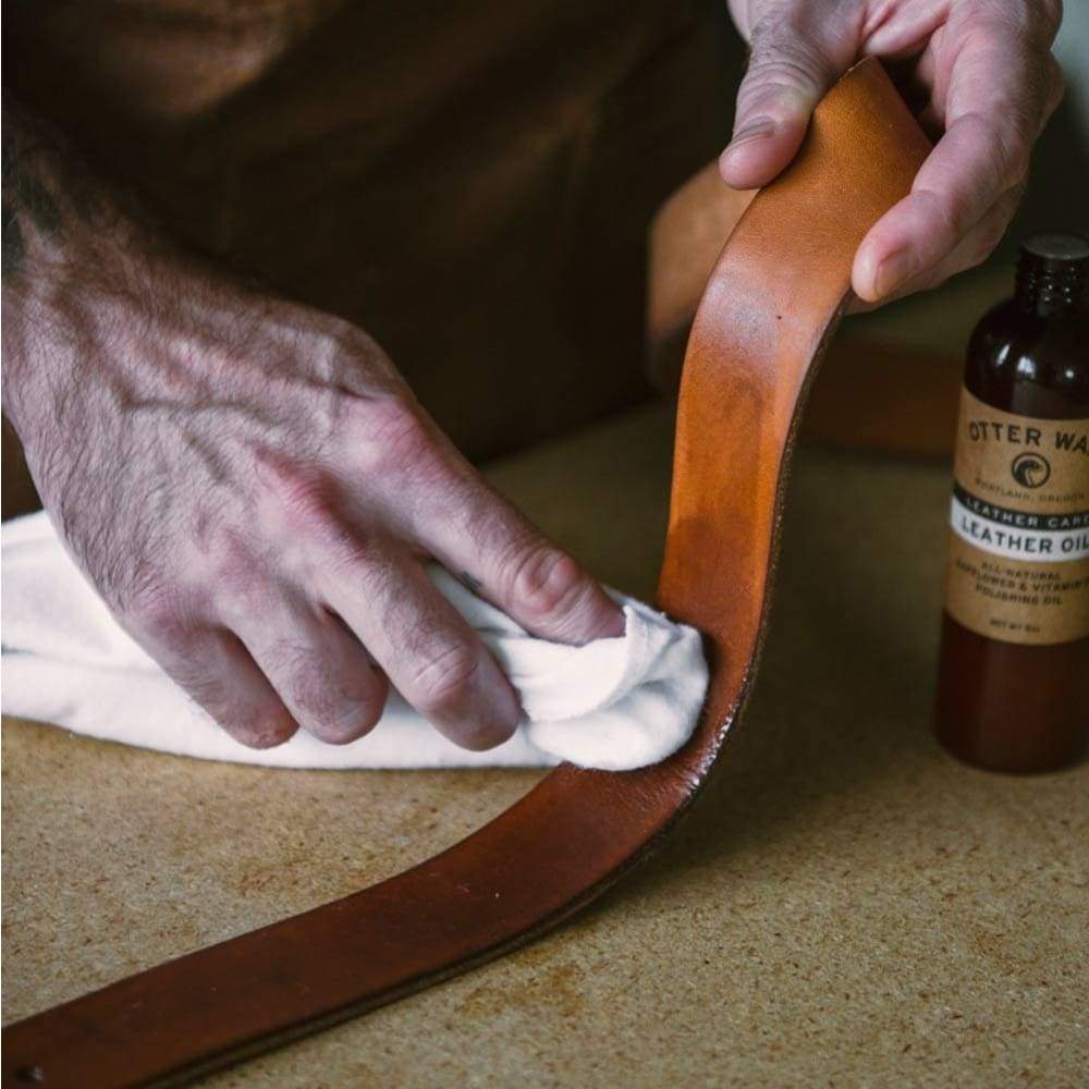 A person uses the Otter Wax Flannel Buffing Cloth to apply leather oil to a brown leather belt. A bottle labeled "Leather Oil" and the Otter Wax cloth are on the table.