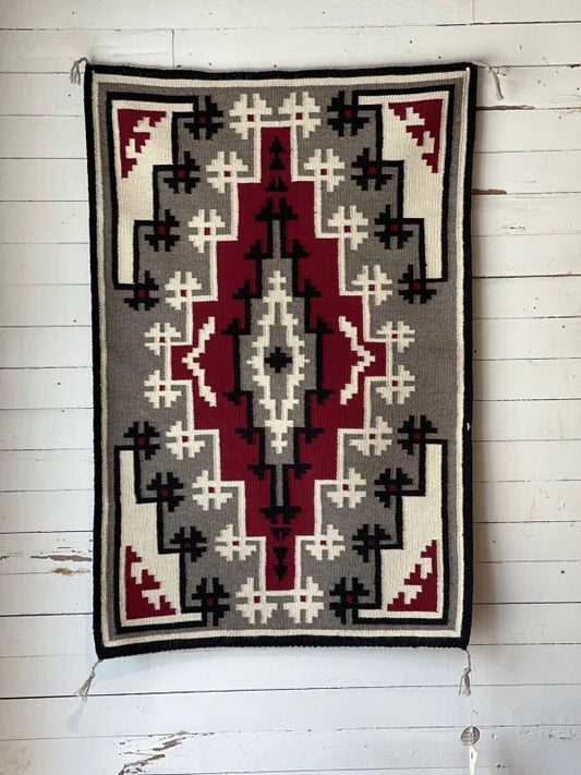 The Ganado Rug Classic "Snowflake" CA 1980 by Vintage, a rectangular Navajo rug featuring red, black, white, and gray geometric patterns in the Ganado style, hangs on a white wooden wall.