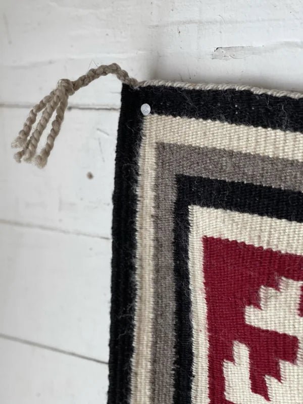 Close-up of a corner of the Vintage Ganado Rug Classic "Snowflake" CA 1980, featuring black, gray, white, and red geometric patterns, attached to a white wooden wall with a nail.