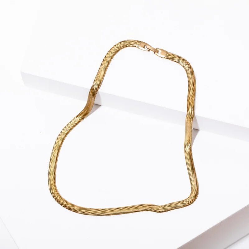 The Grace Necklace by Larissa Loden, a gold herringbone chain, is displayed on a white geometric background.