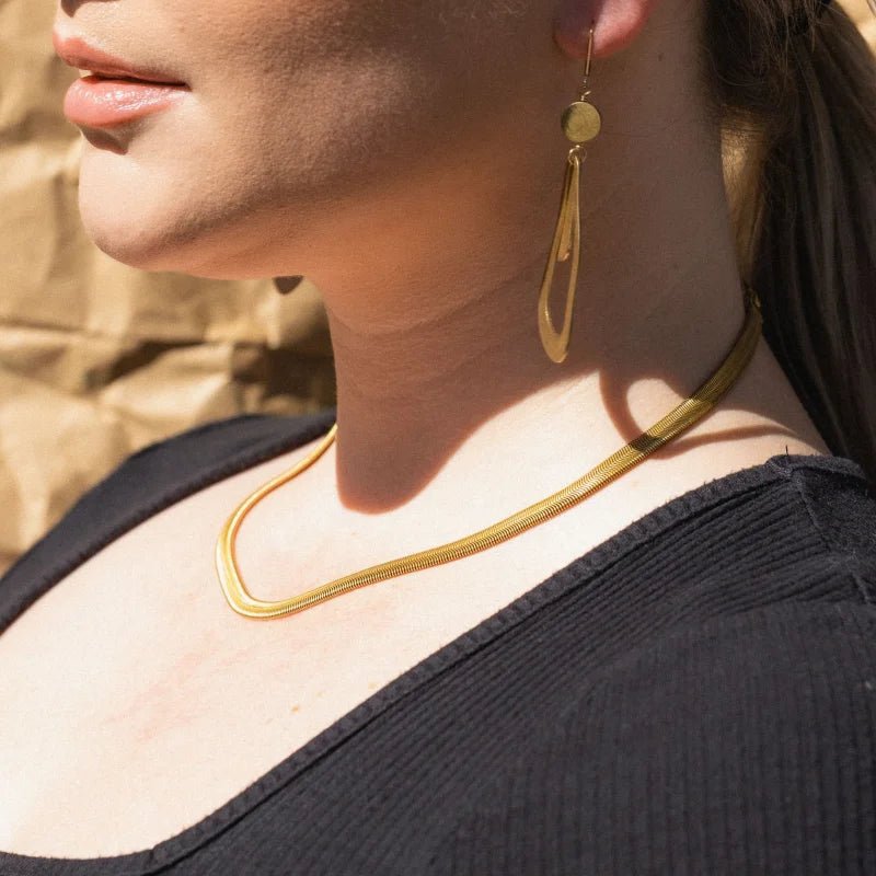 Close-up of a woman wearing the Grace Necklace by Larissa Loden, styled with a long gold earring and a black top against a blurred brown background.
