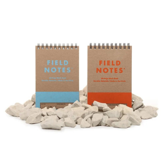 Two upright Field Notes Heavy Duty 2-Pack spiral notebooks, featuring blue and orange text, are displayed on a pile of white rocks against a white background.