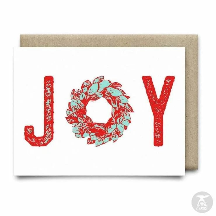 The Joy Wreath Card by Anvil Cards features "JOY" in red with a green wreath "O," printed on premium linen paper. This festive A2 card is blank inside for your personal message.