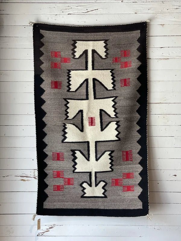 A Vintage Klagetoh Rug Circa 1950 with geometric black, white, gray, and red patterns hangs on a white wooden wall.