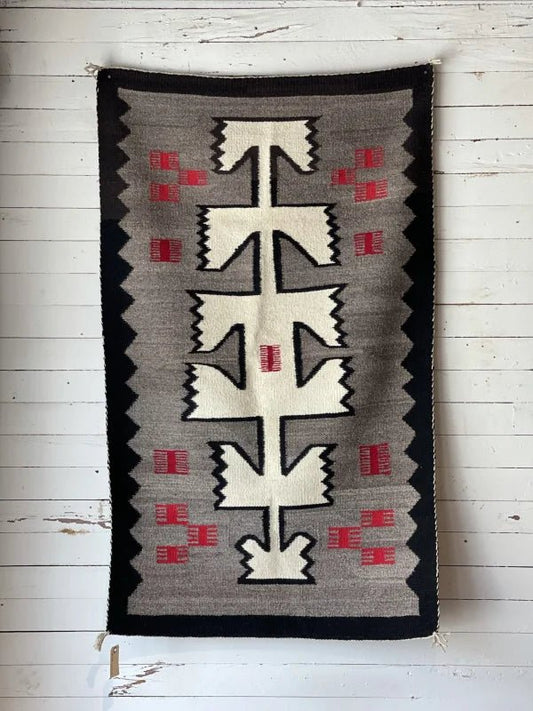 A Vintage Klagetoh Rug Circa 1950 with geometric black, white, gray, and red patterns hangs on a white wooden wall.