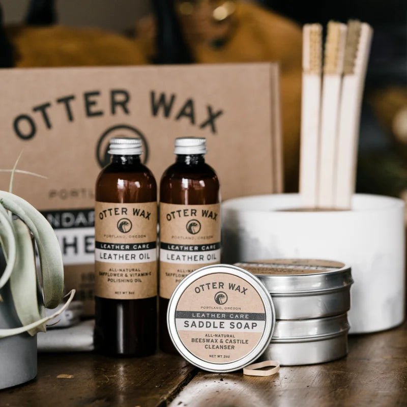 An assortment of Otter Wax leather care products, including Leather Polishing Oil by Otter Wax, saddle soap tins, wooden brushes, and a small potted plant, is displayed neatly on a table.