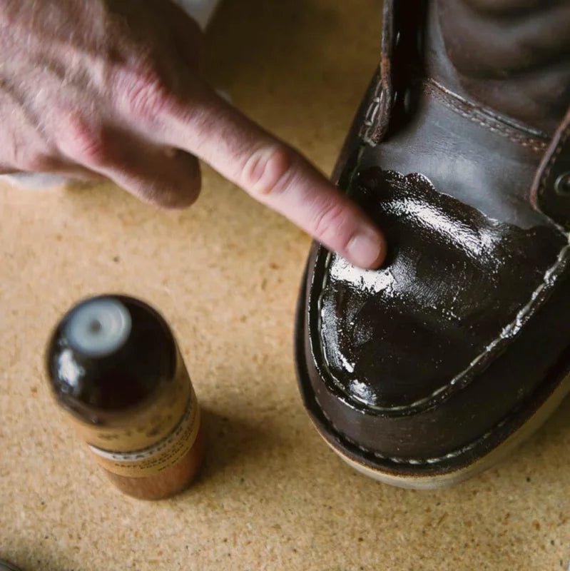 Someone applies Otter Wax Leather Polishing Oil to the toe of a brown leather shoe with their finger, while a bottle of Otter Wax all-natural leather care sits nearby on a wooden surface.
