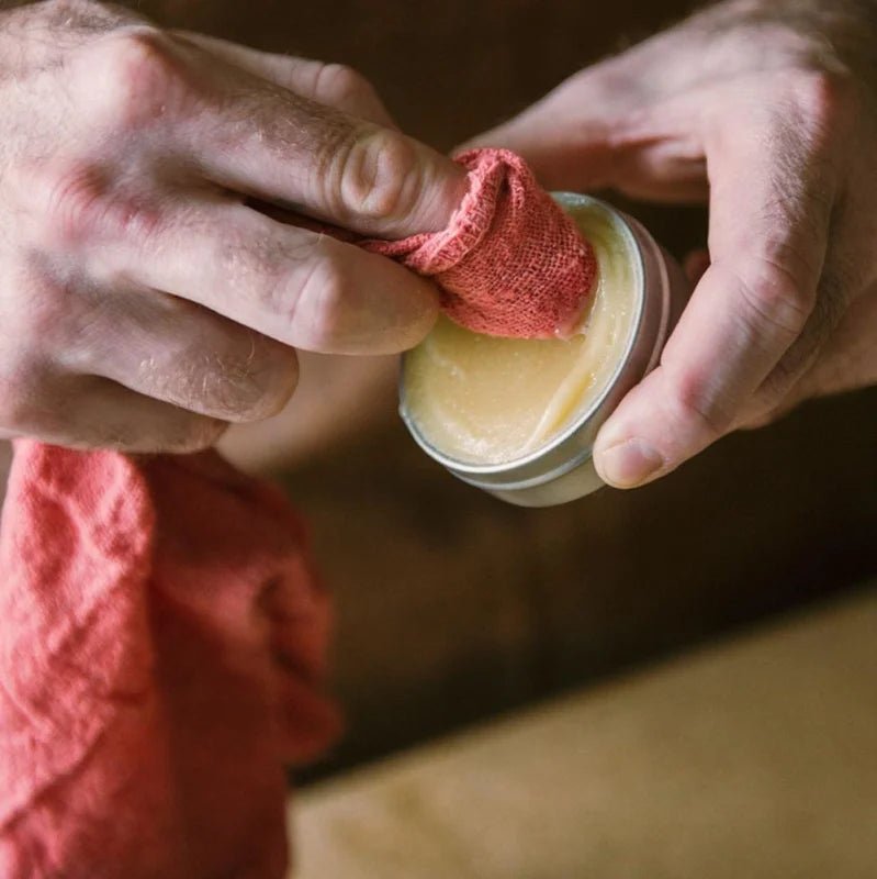 A person uses a red cloth to apply Otter Wax Leather Salve from a small metal tin, deeply conditioning the leather.