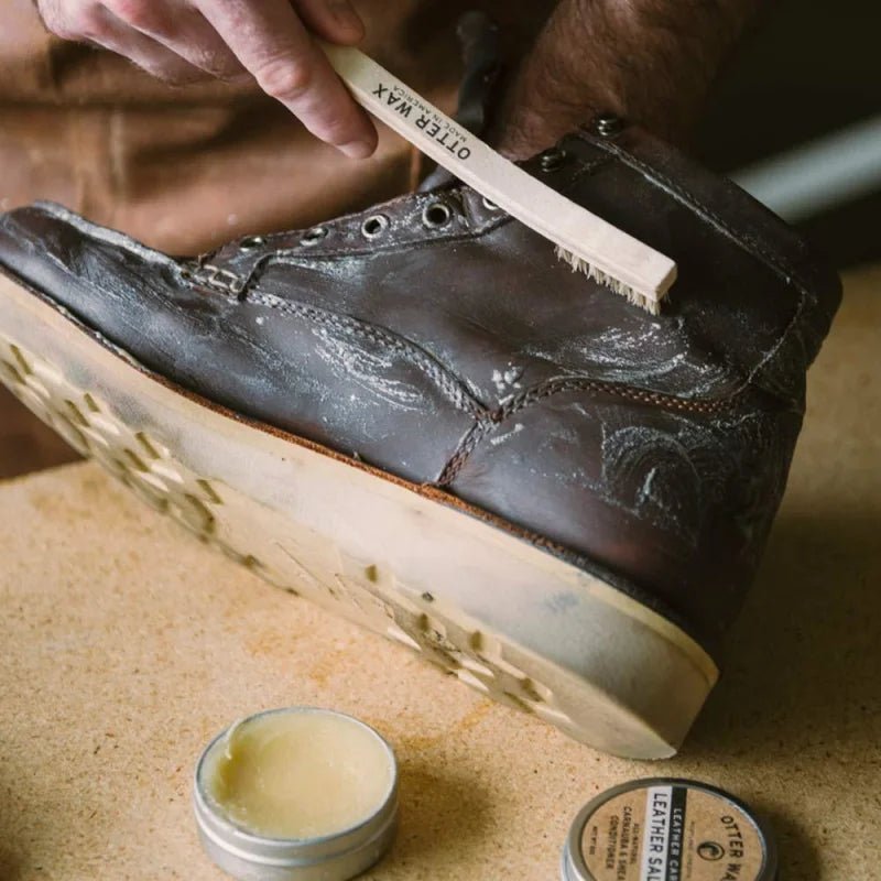 A person brushes a worn brown leather boot with a wooden brush near an open tin of Otter Wax Leather Salve and other Otter Wax leather care products on a table.