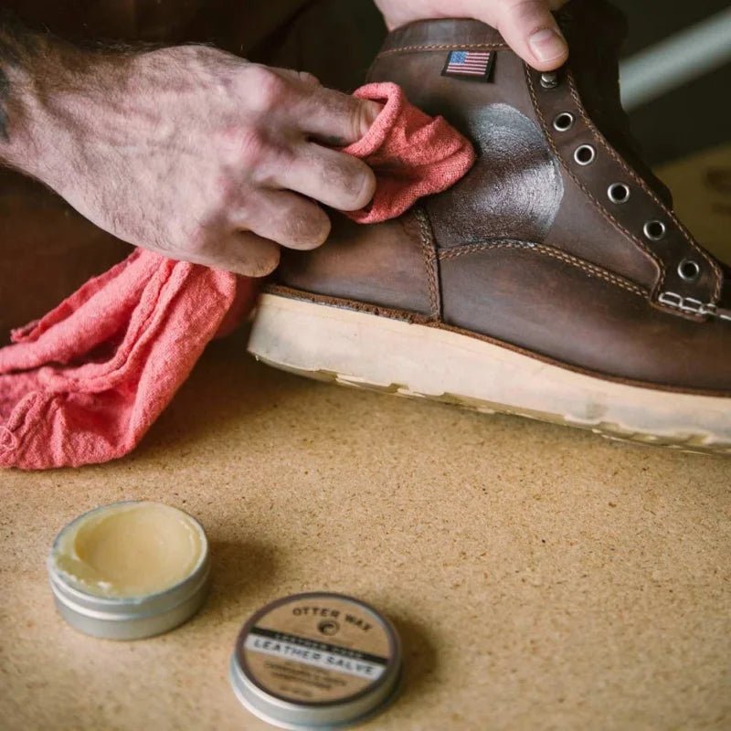 A person polishes a brown leather boot with a red cloth; an open tin of Otter Wax Leather Salve sits on the table nearby.