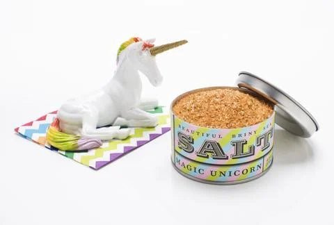 A tin of Beautiful Briny Sea's "Magic Unicorn Sea Salt," lid off to reveal the blend, sits beside a white unicorn figurine on a vibrant zigzag-patterned napkin.