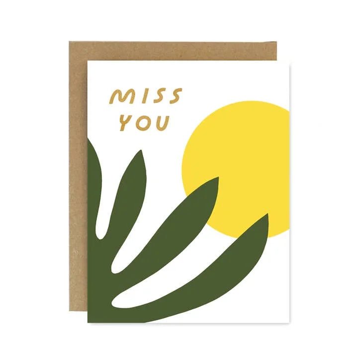 The Miss You Shapes & Colors Card by Worthwhile Paper features green leaves, a bold yellow sun, and "MISS YOU" text on recycled paper. Comes with a brown envelope—an eco-friendly way to send your message.