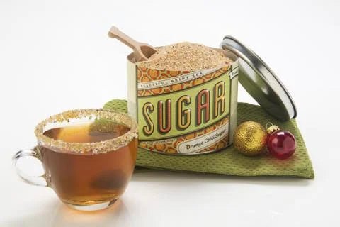 A glass cup of tea with a sugared rim sits beside an open tin of Beautiful Briny Sea's Orange Chili Sugar and two Christmas ornaments on a green cloth.