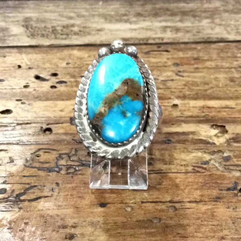 The Vintage Oval Turquoise w/ Twisted Edge Ring, size 5 sterling silver with a large oval stone and bold brown veining, is shown on a clear stand atop a wooden surface.