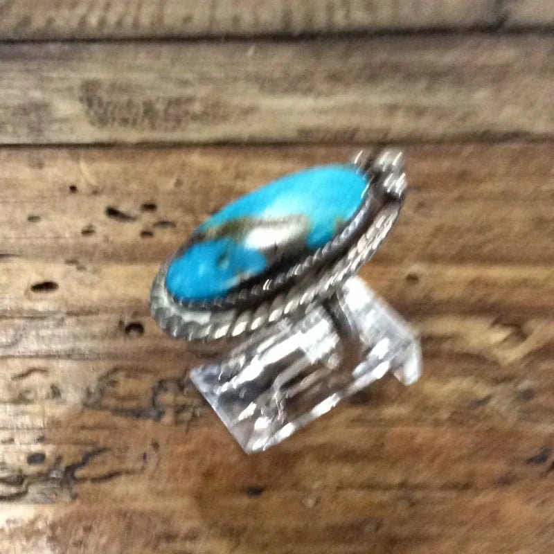 The Vintage Oval Turquoise w/ Twisted Edge Ring in size 5, featuring a large oval turquoise stone, is displayed on a clear stand atop a rustic wooden surface.