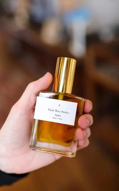 A hand holds a glass perfume bottle with a gold cap and a white label reading "Saint Rita Parlor." The featured unisex scent is Parfum | Signature by Saint Rita Parlor, shown in focus against a blurred background.