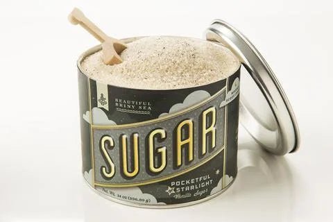 A metal tin labeled "Pocketful of Starlight Vanilla Sugar" by Beautiful Briny Sea is filled with organic cane sugar and comes with a small shovel; the lid is placed next to the can.
