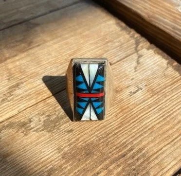 The Vintage Rectangle Multi-stone Ring, size 10 in sterling silver with geometric white, blue, black, and red inlay, rests on a wooden surface in sunlight.