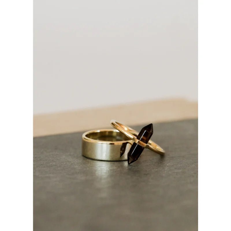 Two gold vermeil rings from JaxKelly rest on a dark surface: one is plain and wide, the other is the hand-crafted Ring | Smokey Quartz Point with a thin band and a horizontally set dark quartz stone.