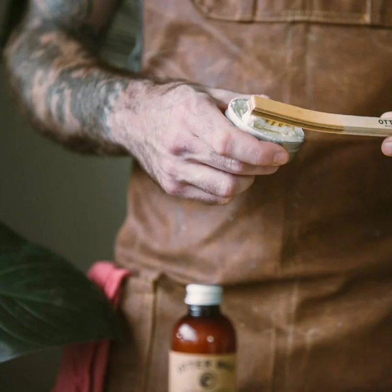 Wearing a brown apron, someone uses a wooden brush to apply Otter Wax Saddle Soap from a small tin, with an Otter Wax bottle and another saddle soap on the table in front of them.