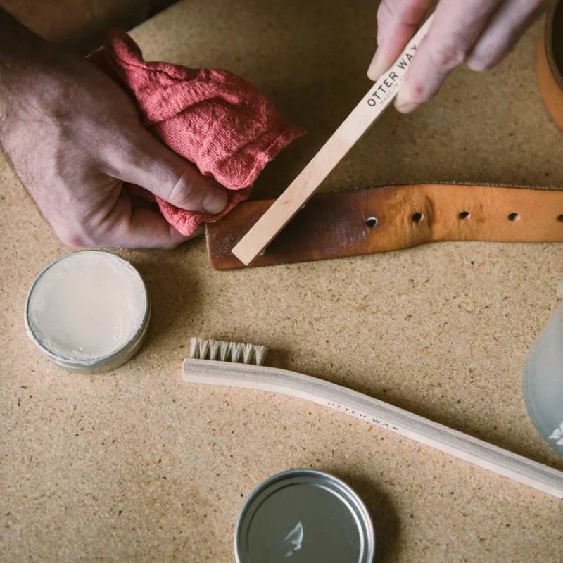 A person cleans a leather belt with a cloth and Otter Wax all-natural care products. On the table are Otter Wax Saddle Soap, a tin of balm, a brush, and a wooden tool labeled "OTTER WAX.
