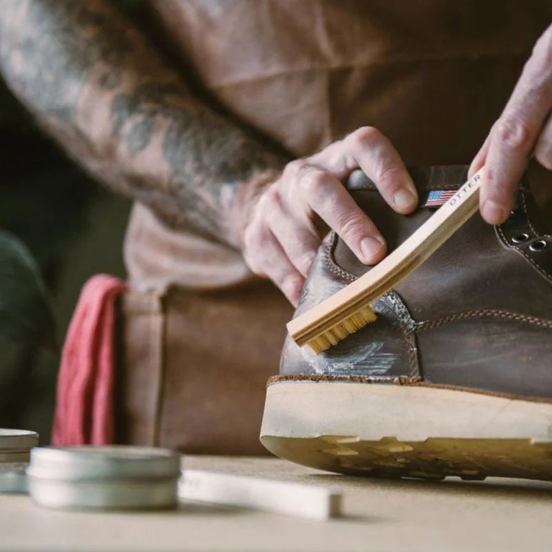 A person uses a small brush to apply all-natural conditioner to a brown leather boot, with Otter Wax Saddle Soap and other Otter Wax shoe care products on the table.