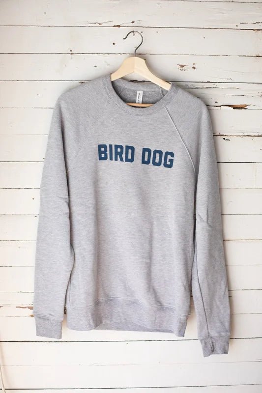 The Ballad of the Bird Dog "BIRD DOG" Shop Sweater is a gray unisex crewneck made in the USA, featuring bold blue text on the chest. It hangs on a wooden hanger against a white wooden wall.