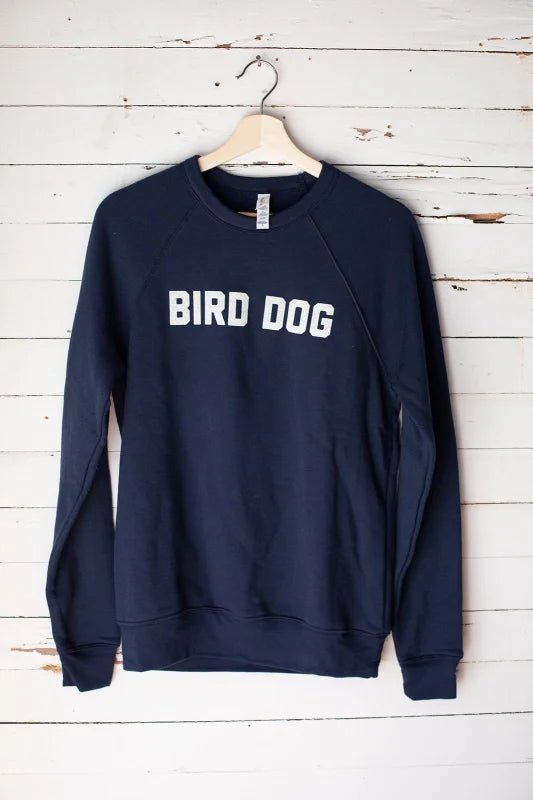 The Bird Dog Sweater by Ballad of the Bird Dog is a navy blue unisex crewneck with "BIRD DOG" in white on the front, made in the USA, shown on a wooden hanger against a white wood wall.