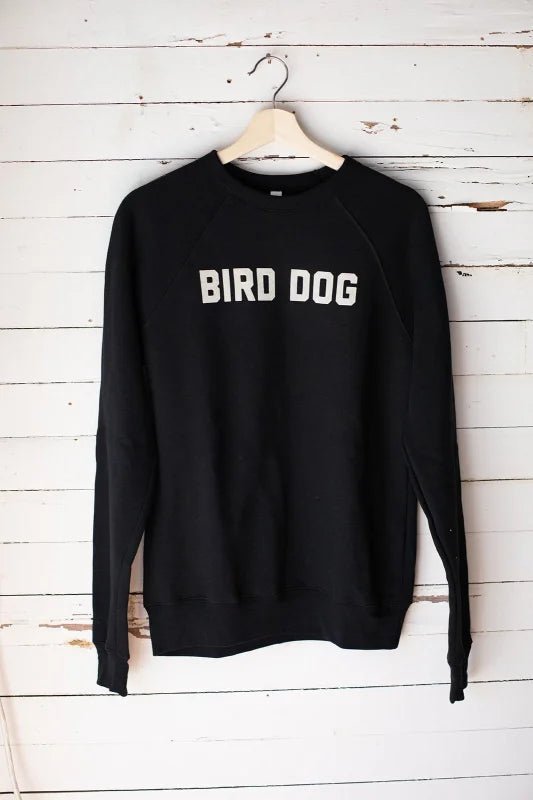 The Ballad of the Bird Dog “Shop Sweater”—a unisex crewneck featuring “BIRD DOG” in white—hangs on a wooden hanger against a white wooden wall.