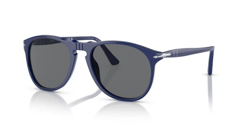 The Persol 0PO9649S Solid Blue w/ Dark Grey sunglasses feature a slim, round unisex design with dark lenses, a keyhole bridge, and silver temple accents, inspired by classic Persol pilot shades and shown on a white background.