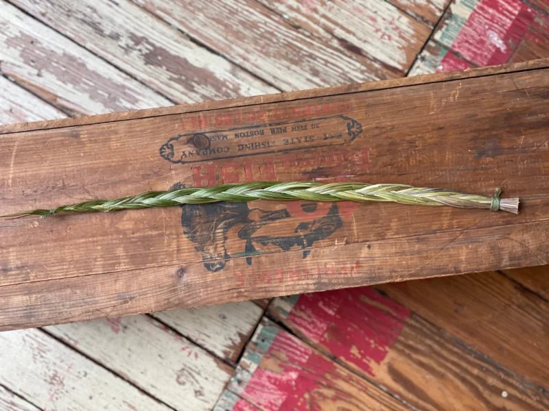 A single long, lush green Sweetgrass braid from Taos Herb Co. lies horizontally on a weathered wooden surface with a red and cream painted pattern.
