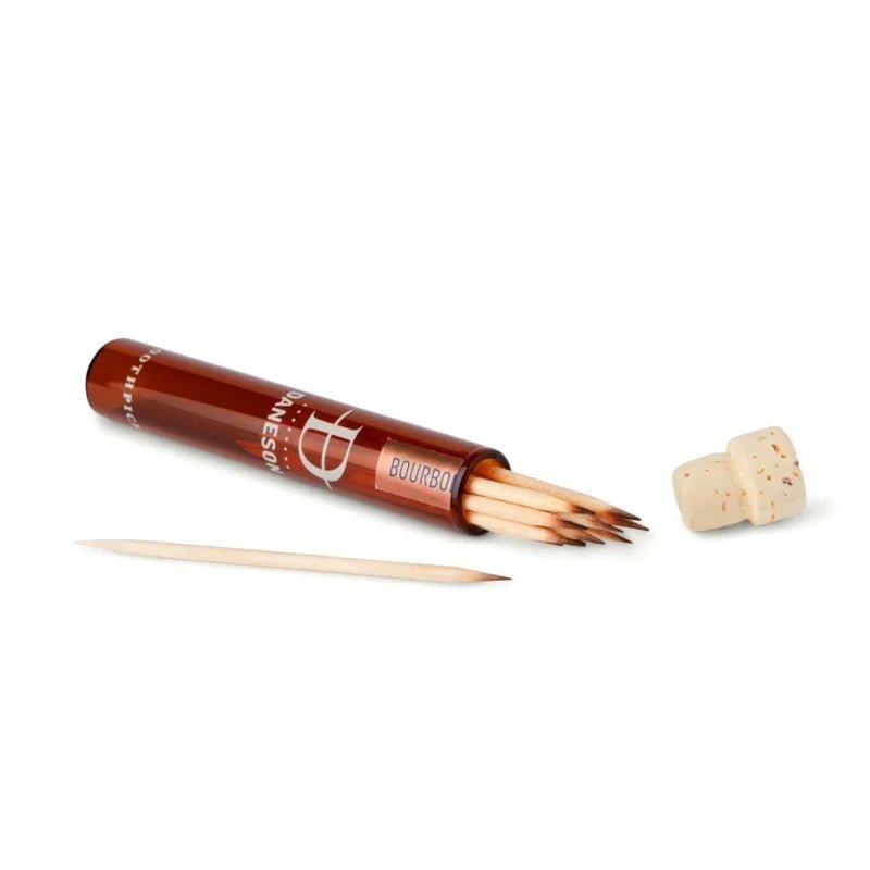 A brown cylindrical Daneson container labeled "Toothpick | Bourbon No. 22" sits open, holding several whiskey-infused toothpicks, with one wooden stick lying beside it.