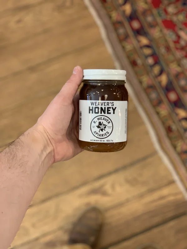 A hand holds a jar of Weaver's Honey by R. Weaver Apiaries with a white label and lid, set on a wooden floor beside a patterned rug—enjoy pure local Navasota honey from R. Weaver Apiaries.