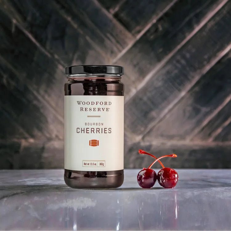 A jar of Woodford Reserve Bourbon Cherries by Bourbon Barrel Foods rests on marble beside two fresh cherriesāan ideal Manhattan cocktail garnishāset against a dark patterned background.