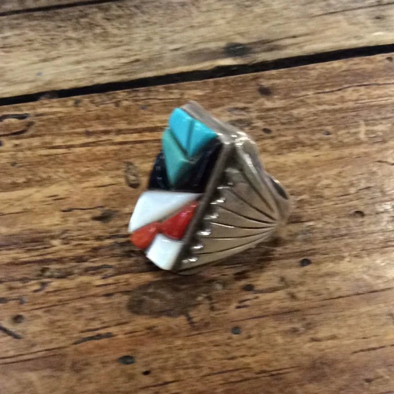 The Zuni Multi-stone Geometric Ring by Vintage showcases inlays of turquoise, red, black, and white stones set in sterling silver, displayed elegantly on a rustic wooden surface.