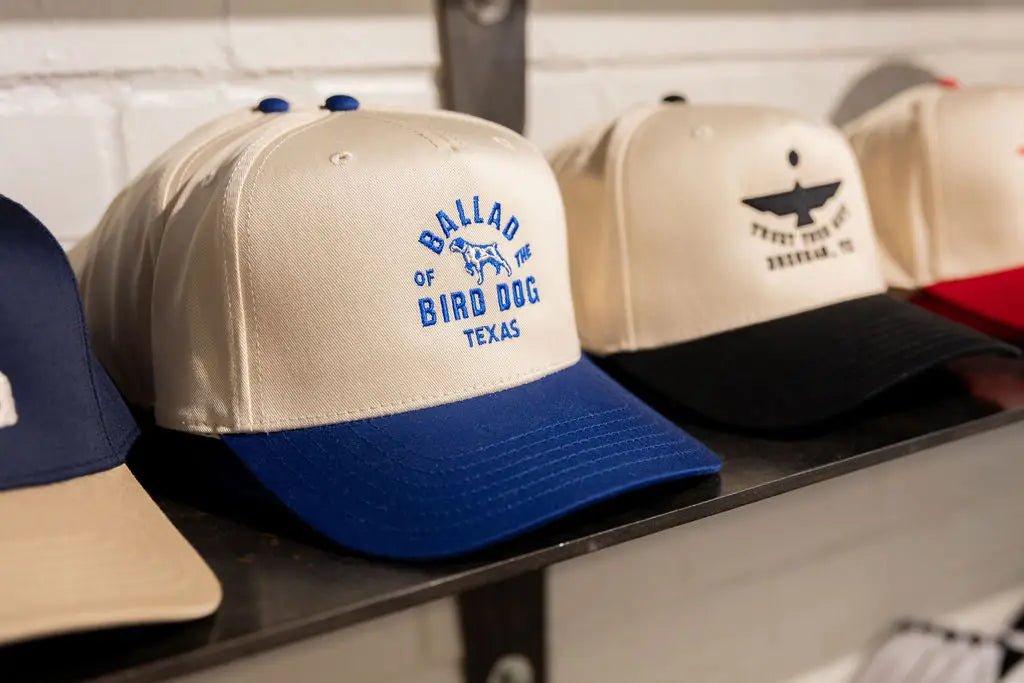 A cream and blue baseball cap on a shelf reads “BALLAD OF BIRD DOG TEXAS” beside other caps in various colors and designs.