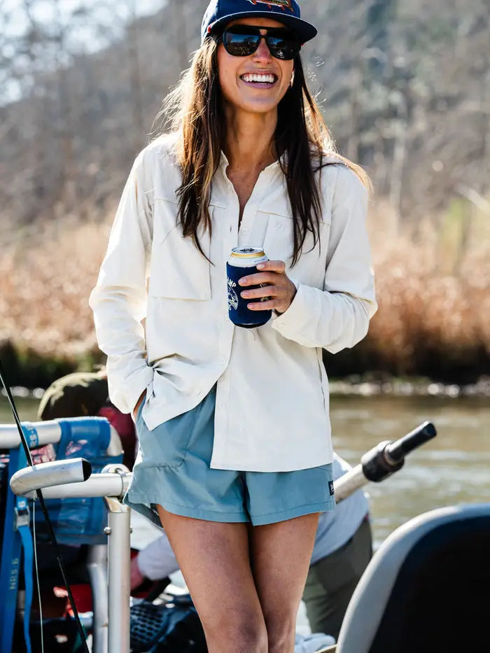 A woman in sunglasses, a cap, and casual outdoor clothing stands smiling by a river, holding a beverage can in a blue holder.