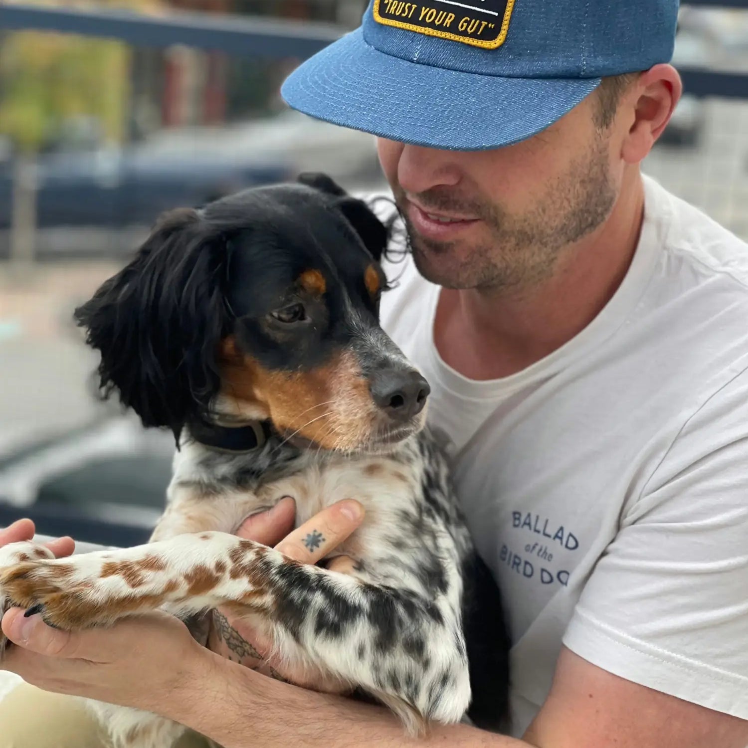 A man wearing a blue "Ballad of the Bird Dog" hat and white shirt holds a black and white dog outside, gently holding its paw.