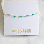 The Turquoise Seed Bead Bracelet by Mesa Blue features a delicate 18K gold plated chain adorned with small turquoise beads and includes an extender chain for the perfect fit, displayed on a white card labeled “Mesa Blue Handcrafted.”.